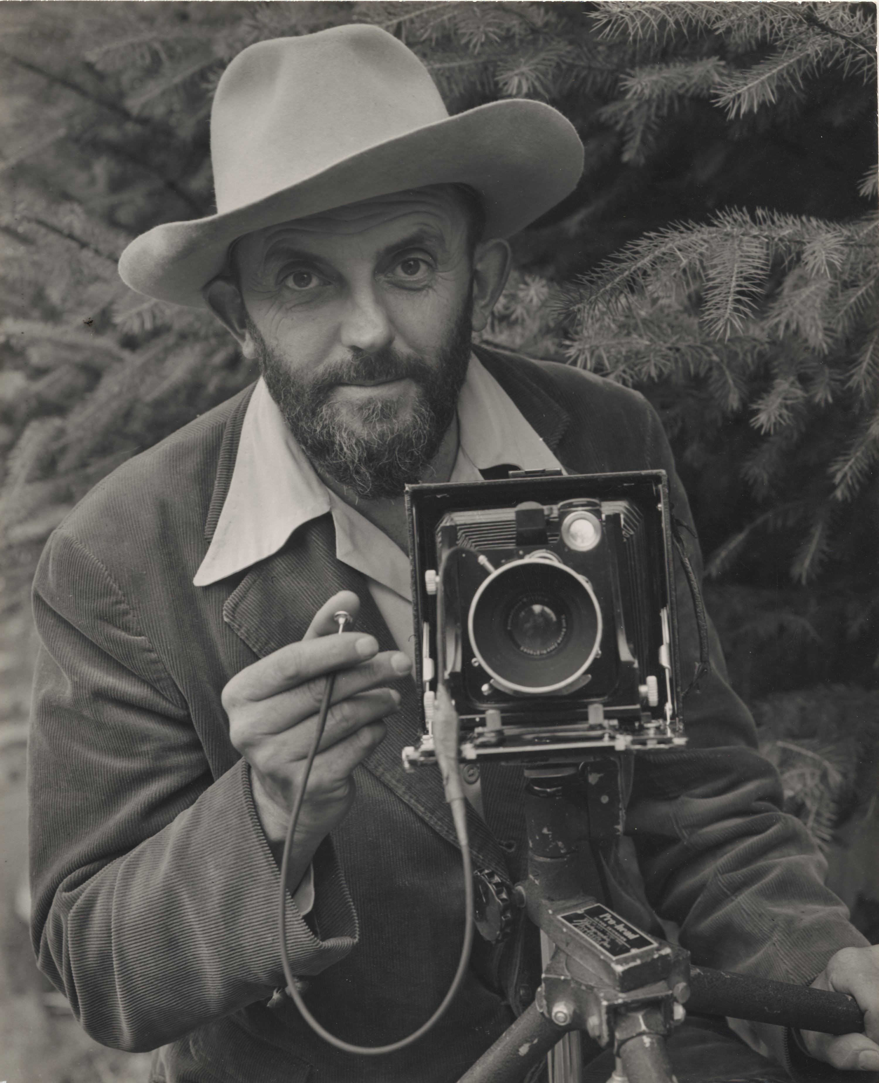 Ansel Adams stands behind a large camera wearing a wide-brimmed hat.
