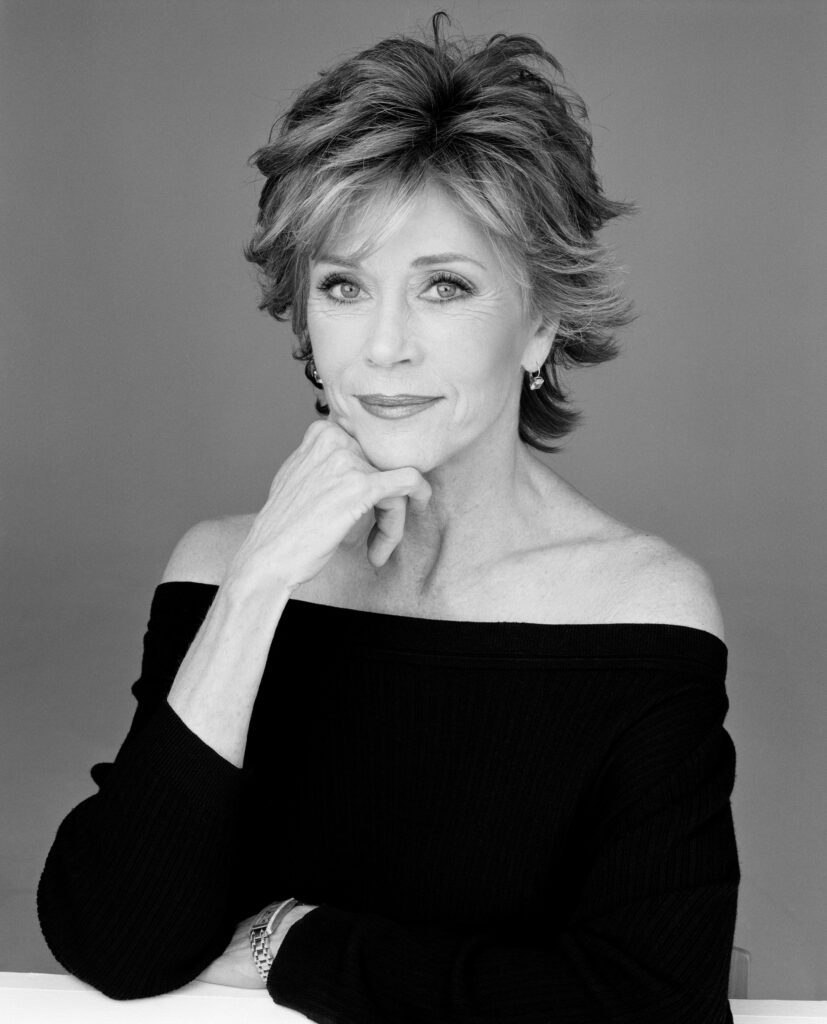 Jane Fonda poses with a hand curled under her cheek and wears a shirt draped off her shoulders.