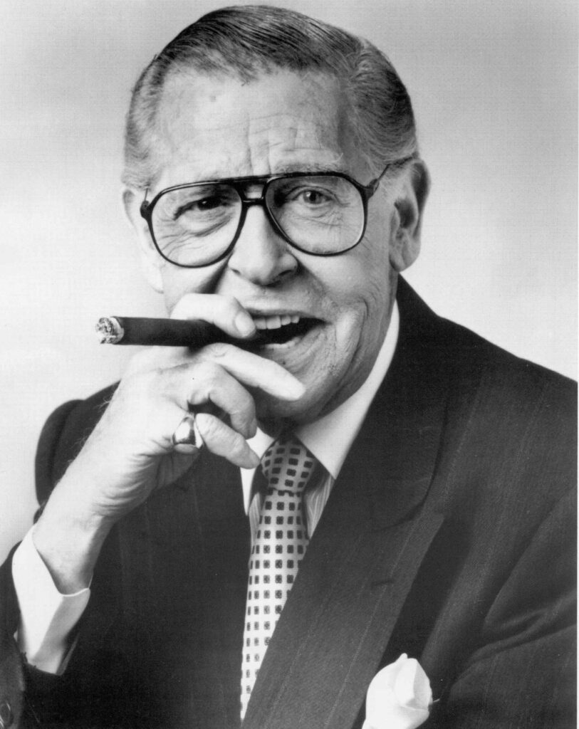 Milton Berle with a cigar in his mouth in a suit and tie.