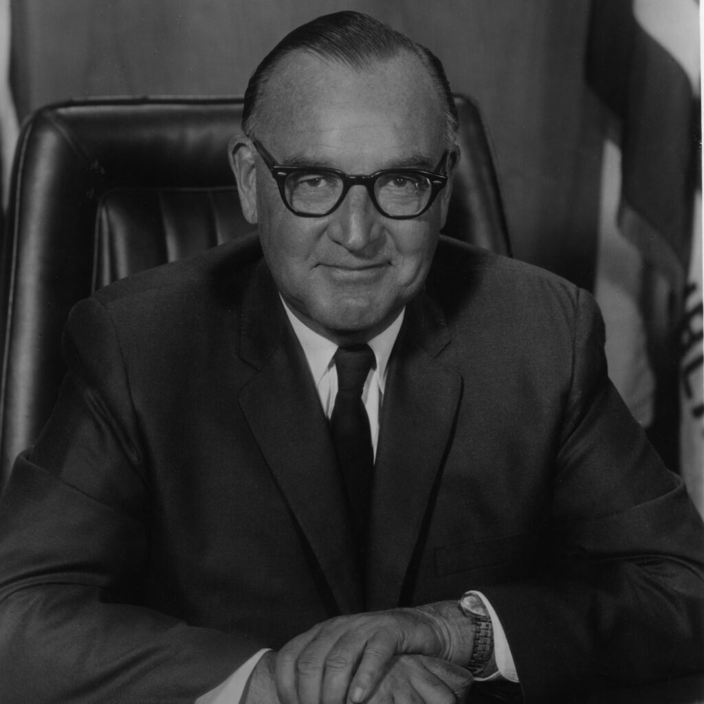 Edmund G. "Pat" Brown sits in a tall leather desk chair wearing a suit and an amused expression.