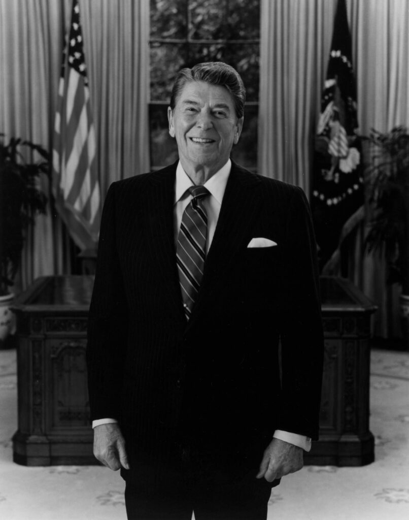 Ronald Reagan stands in front of his Oval Office desk and smiles warmly.
