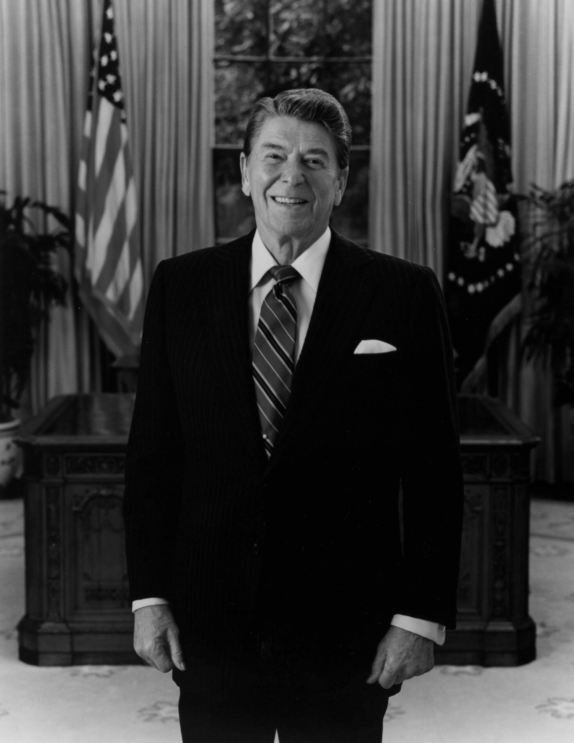 Ronald Reagan stands in front of his Oval Office desk and smiles warmly.