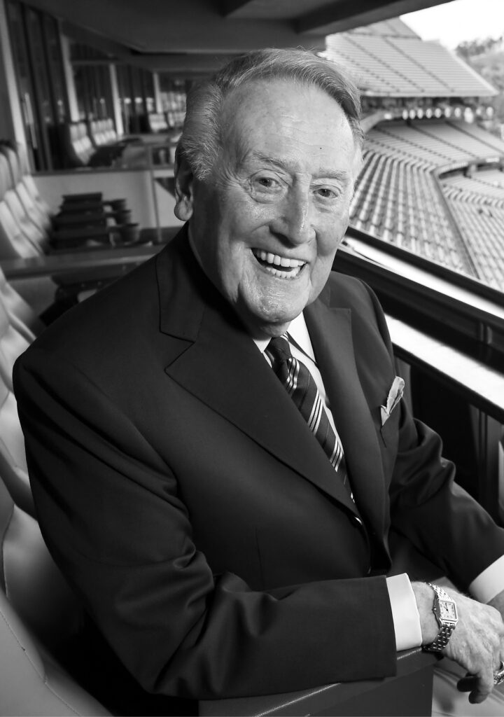 A suited-up Vin Scully sits in Dodger Stadium with a smile.