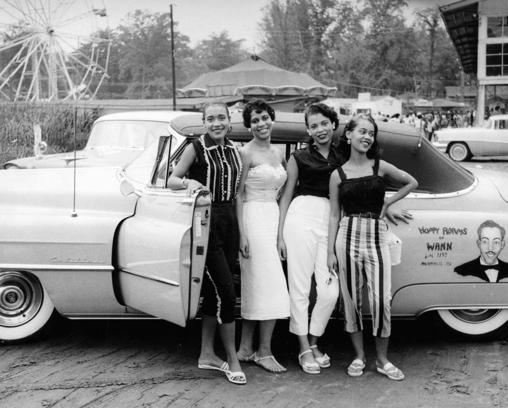 Four women pose in front of a car.