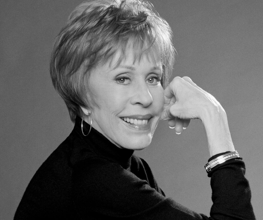 Headshot of a smiling Carol Burnett with fingers touching her cheek.