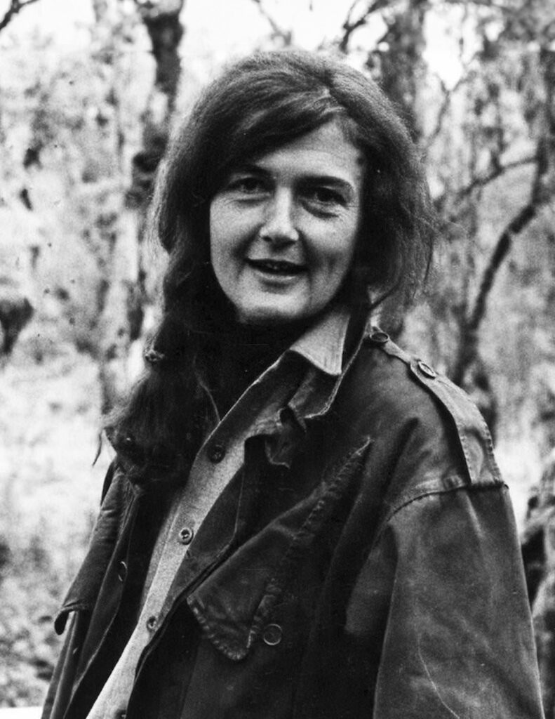 Dian Fossey looking at the camera in the forest, wearing a button up shirt and jacket.