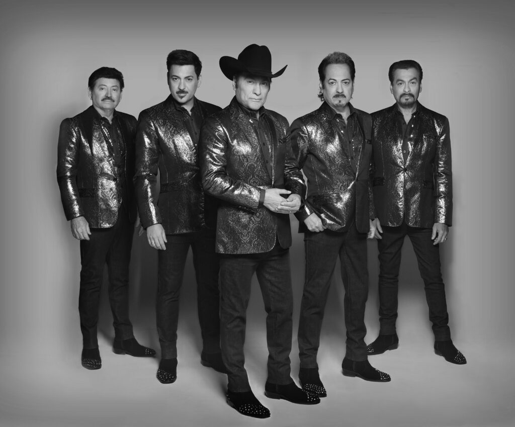 Los Tigres del Norte pose in shiny suits in front of a plain backdrop.