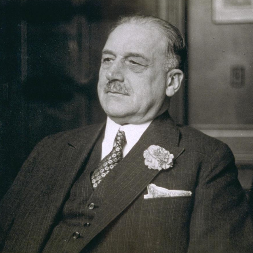 A.P. Giannini wears a suit and tie and leans back with an amused expression.