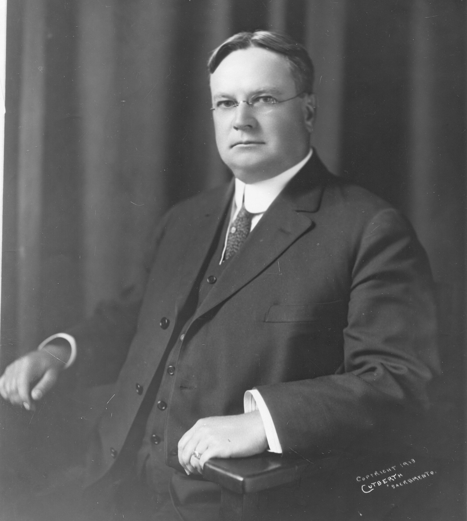 Hiram Johnson sits in a chair with a two-piece suit and a stern expression.
