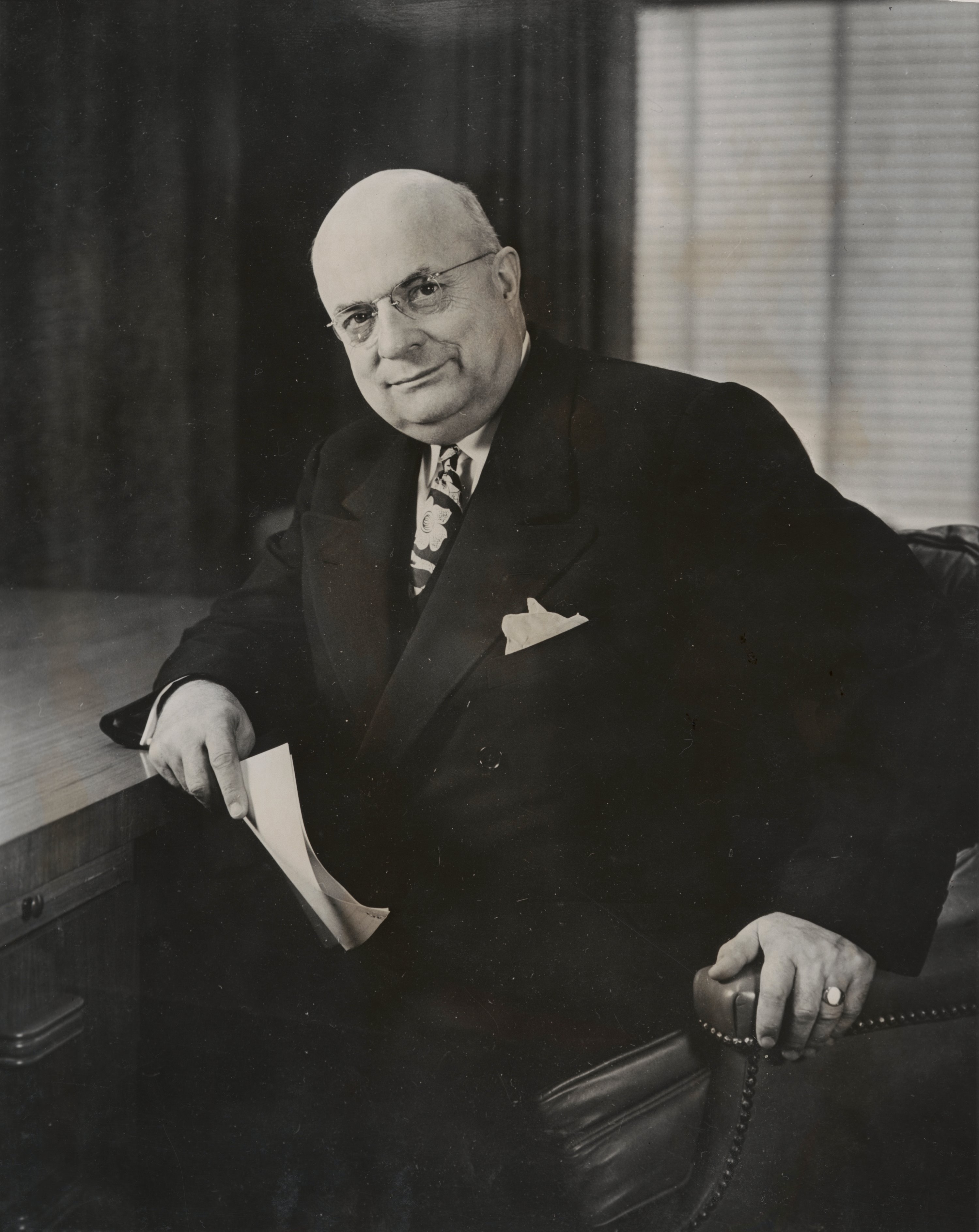 Henry J. Kaiser leans against a desk with papers in hand wearing a suit.