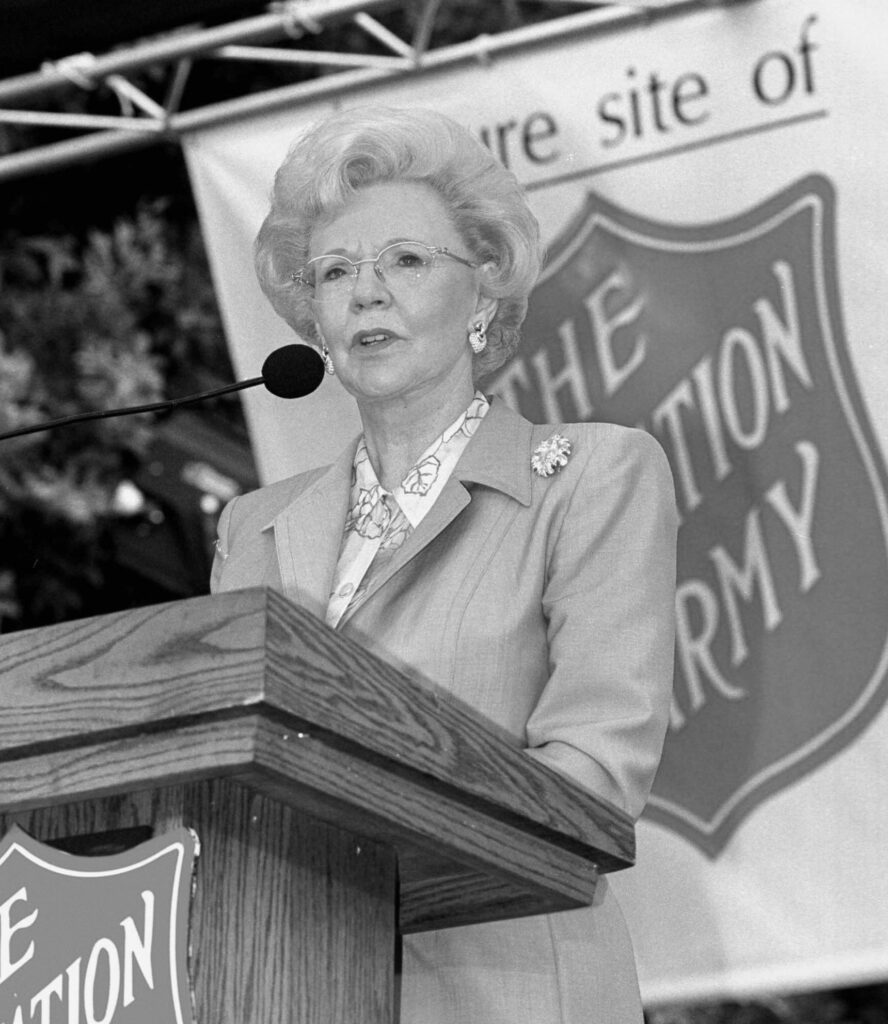 Joan Kroc speaks at a podium with a Salvation Army logo behind her.