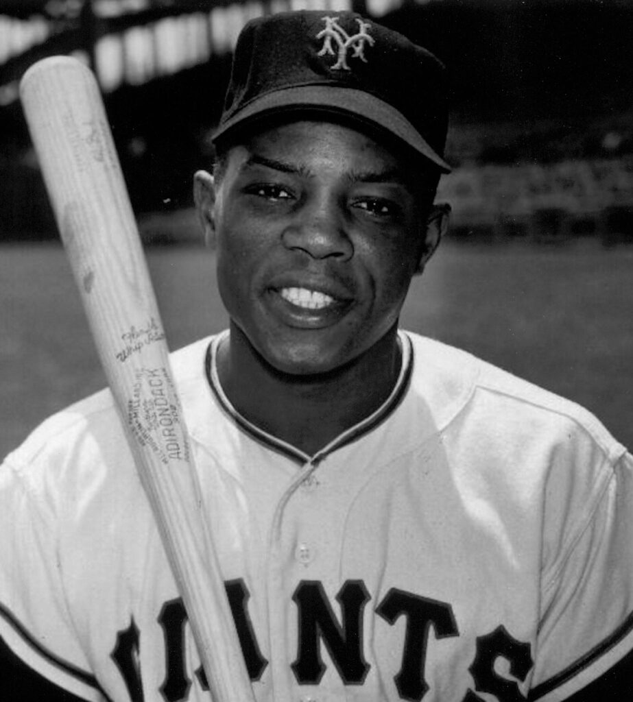 A young Willie Mays poses with his New York Giants cap and jersey and holds a bat.