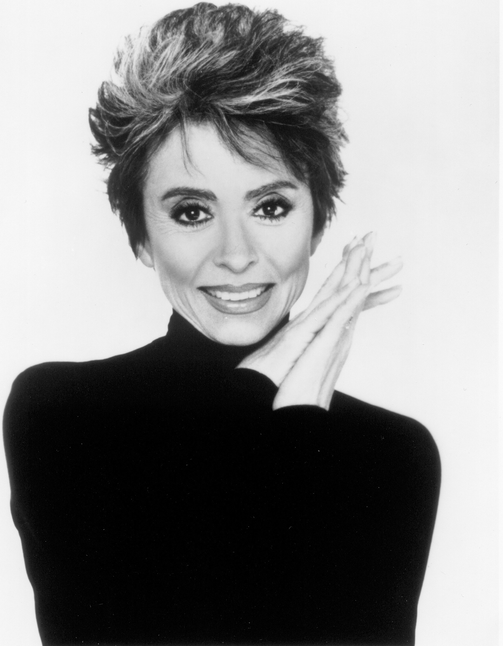 Headshot of a smiling Rita Moreno with hands folded at her face.