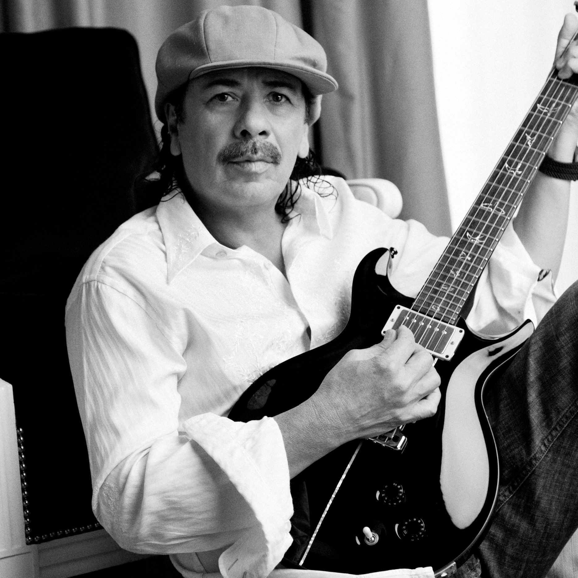 Carlos Santana poses with his guitar wearing a news cap and button-down shirt.