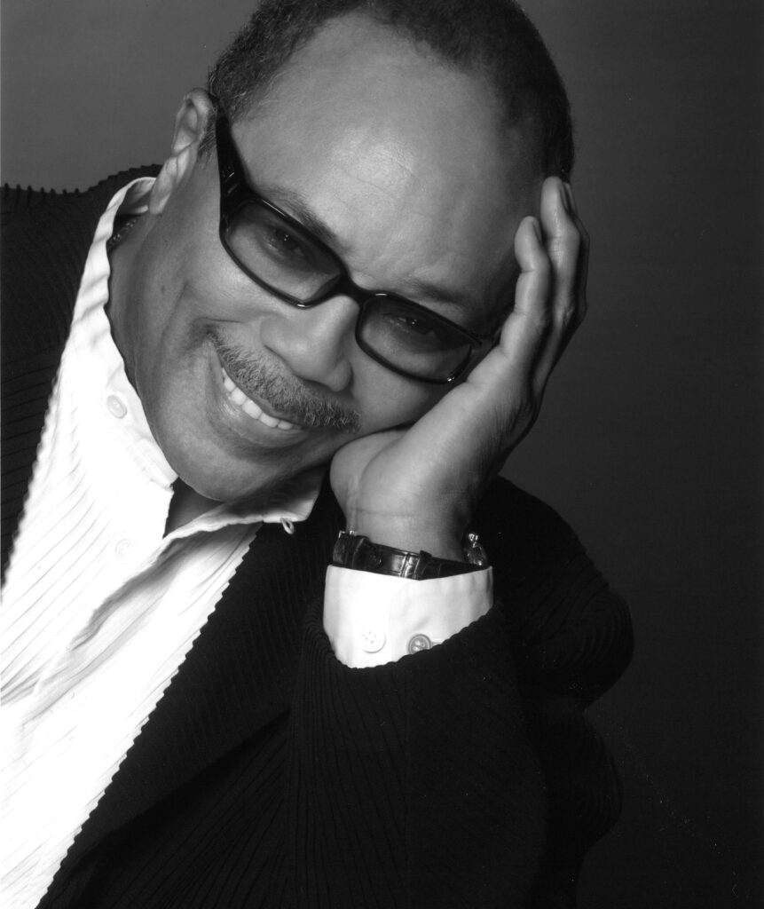 Headshot of a smiling Quincy Jones with thick-rimmed glasses and his head rested in palm.