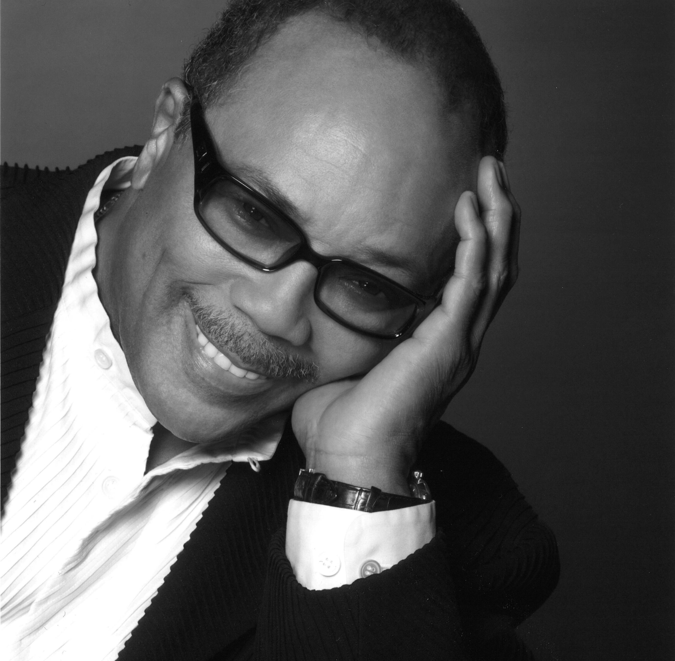 Headshot of a smiling Quincy Jones with thick-rimmed glasses and his head rested in palm.