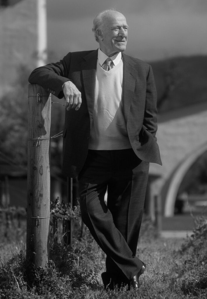 Robert Mondavi leans against the wood post of a wired fence and smiles.