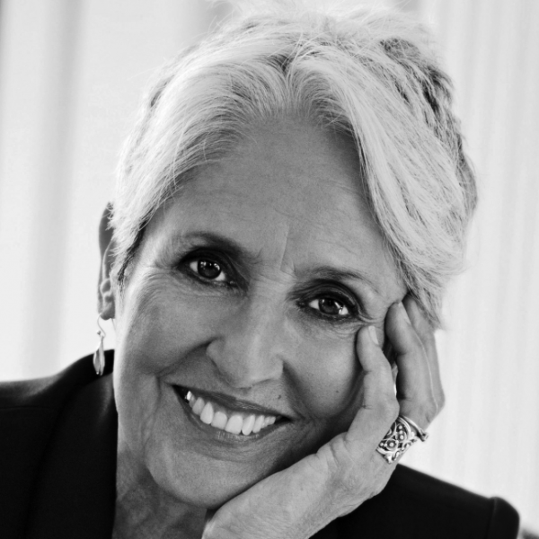 Joan Baez smiles warmly while resting her face against her palm.