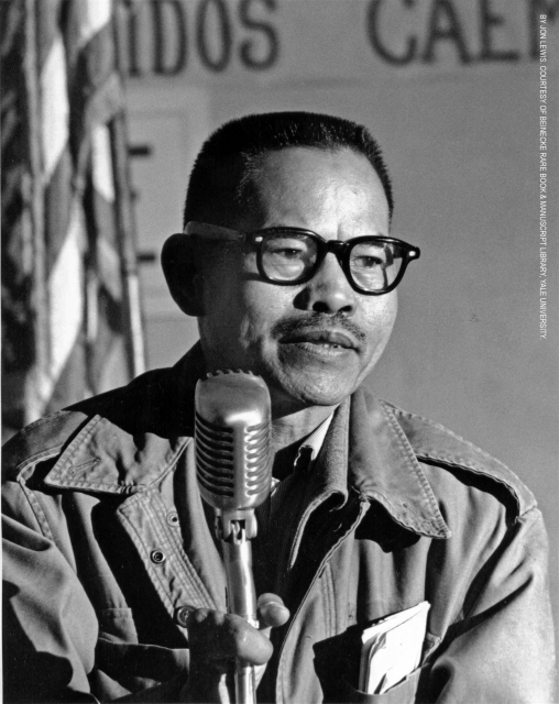 A young Larry Itliong speaks at a standing microphone.