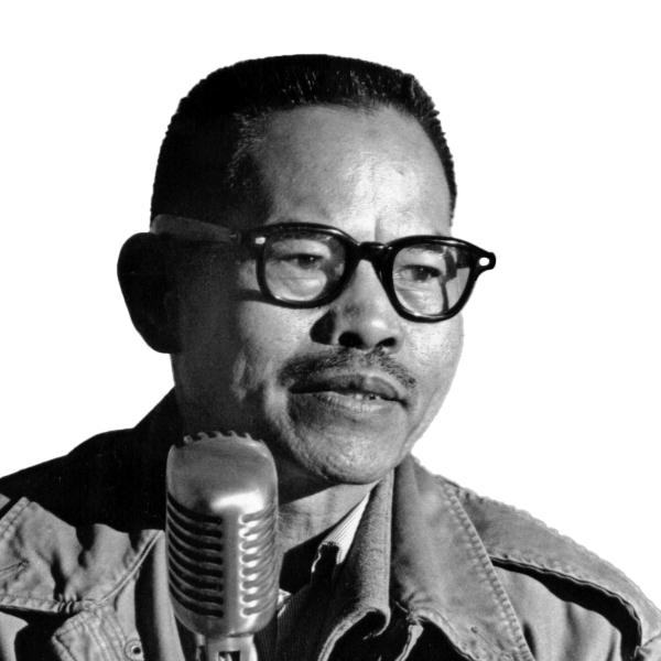 A young Larry Itliong speaks at a standing microphone.