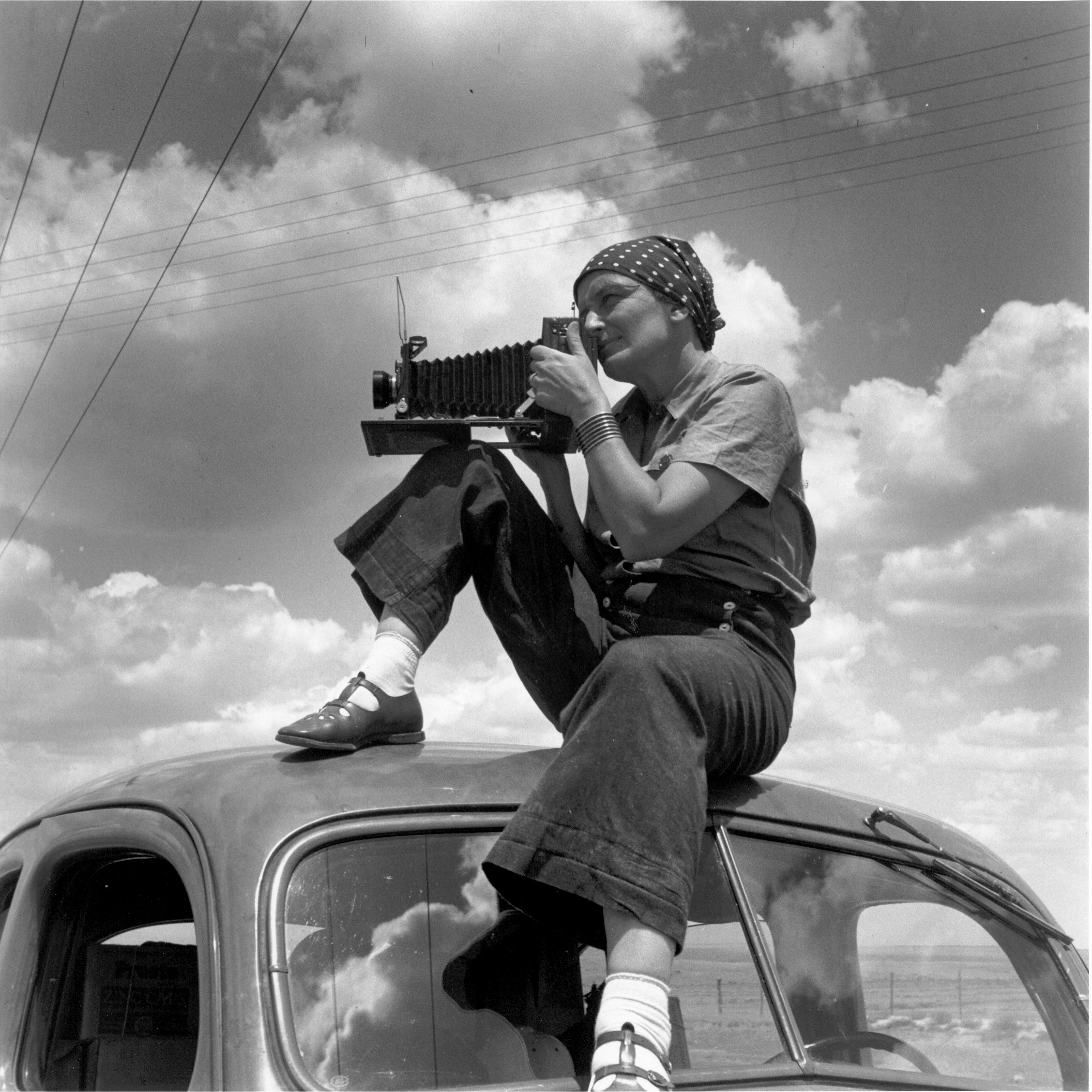 Dorothea Lange sits atop a car and holds her camera to her face.