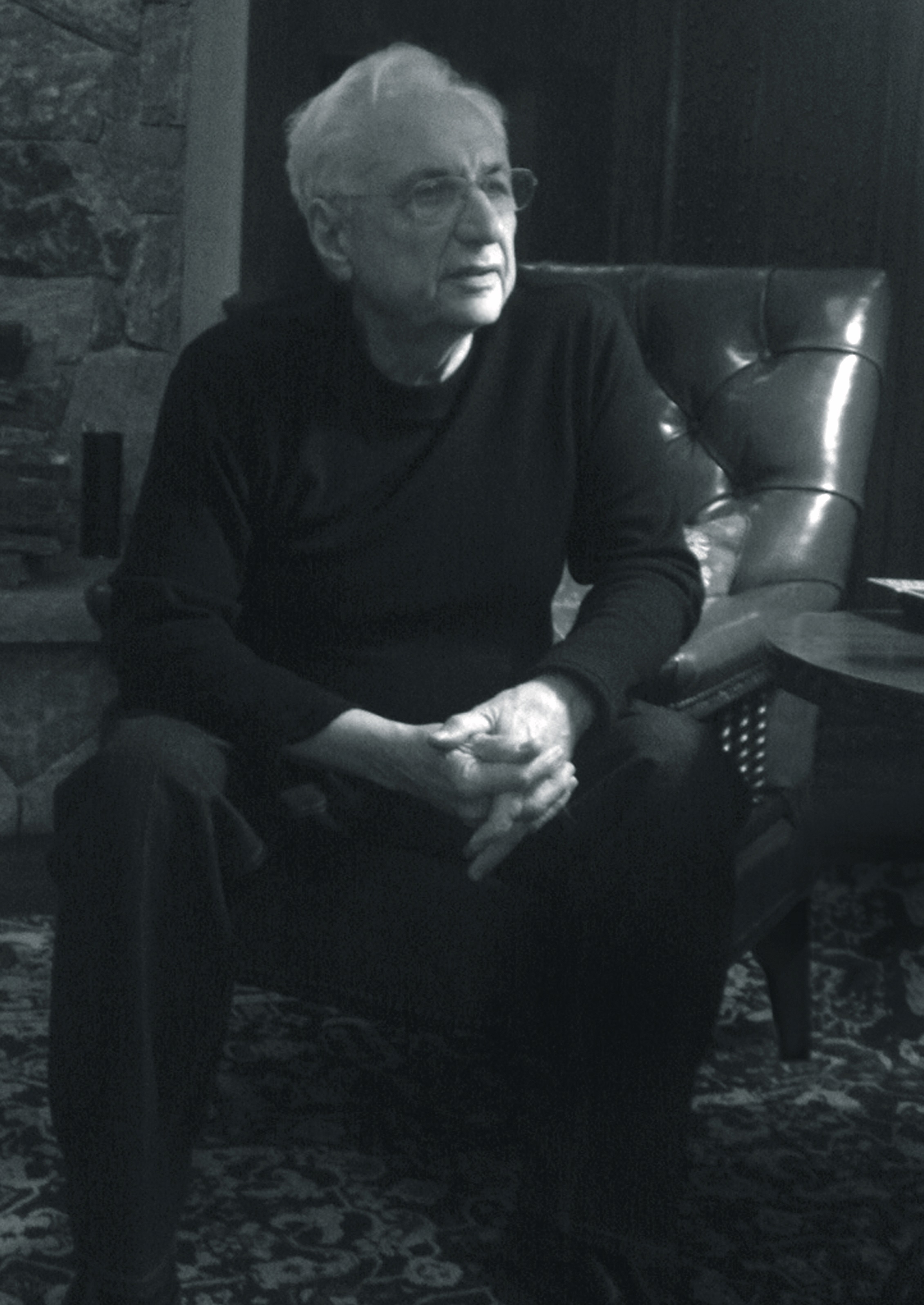 Frank Gehry sits with hands folded and a pondering gaze.