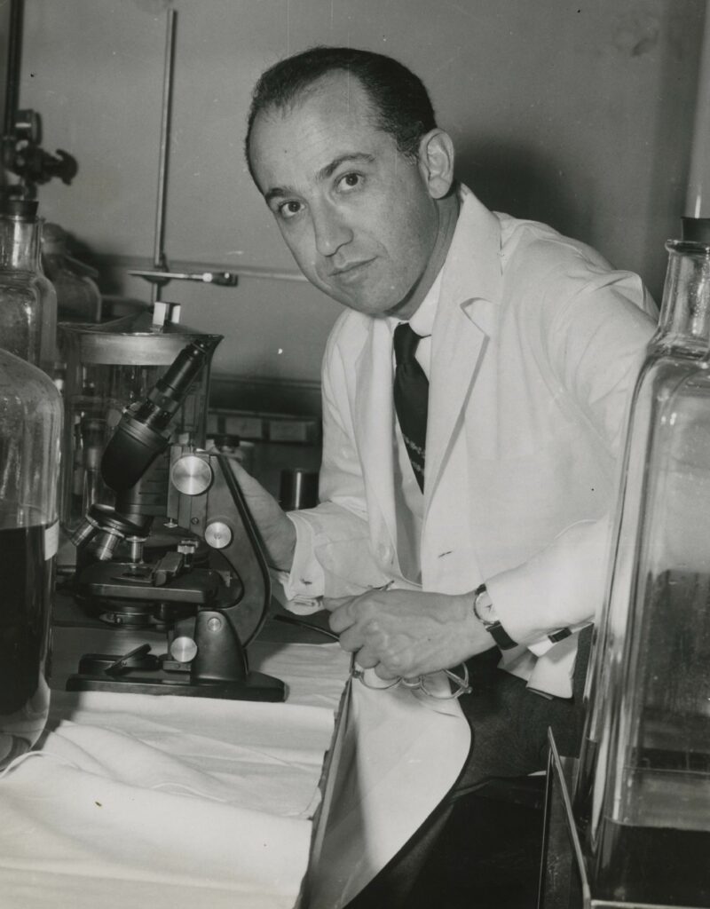 Jonas Salk looks up from his microscope and wears lab attire.