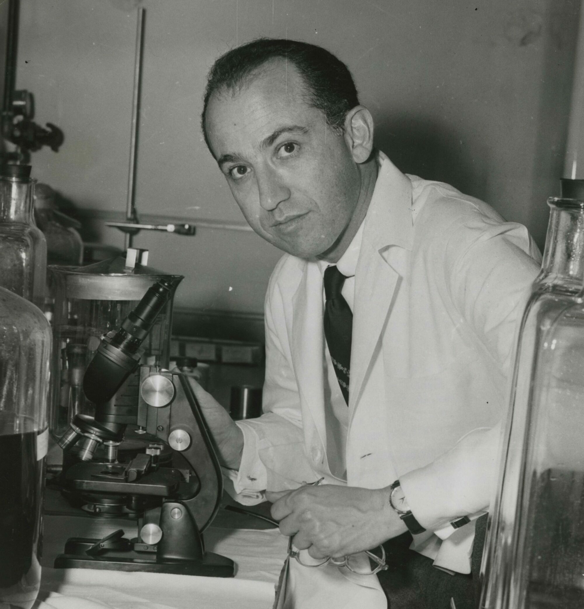 Jonas Salk looks up from his microscope and wears lab attire.