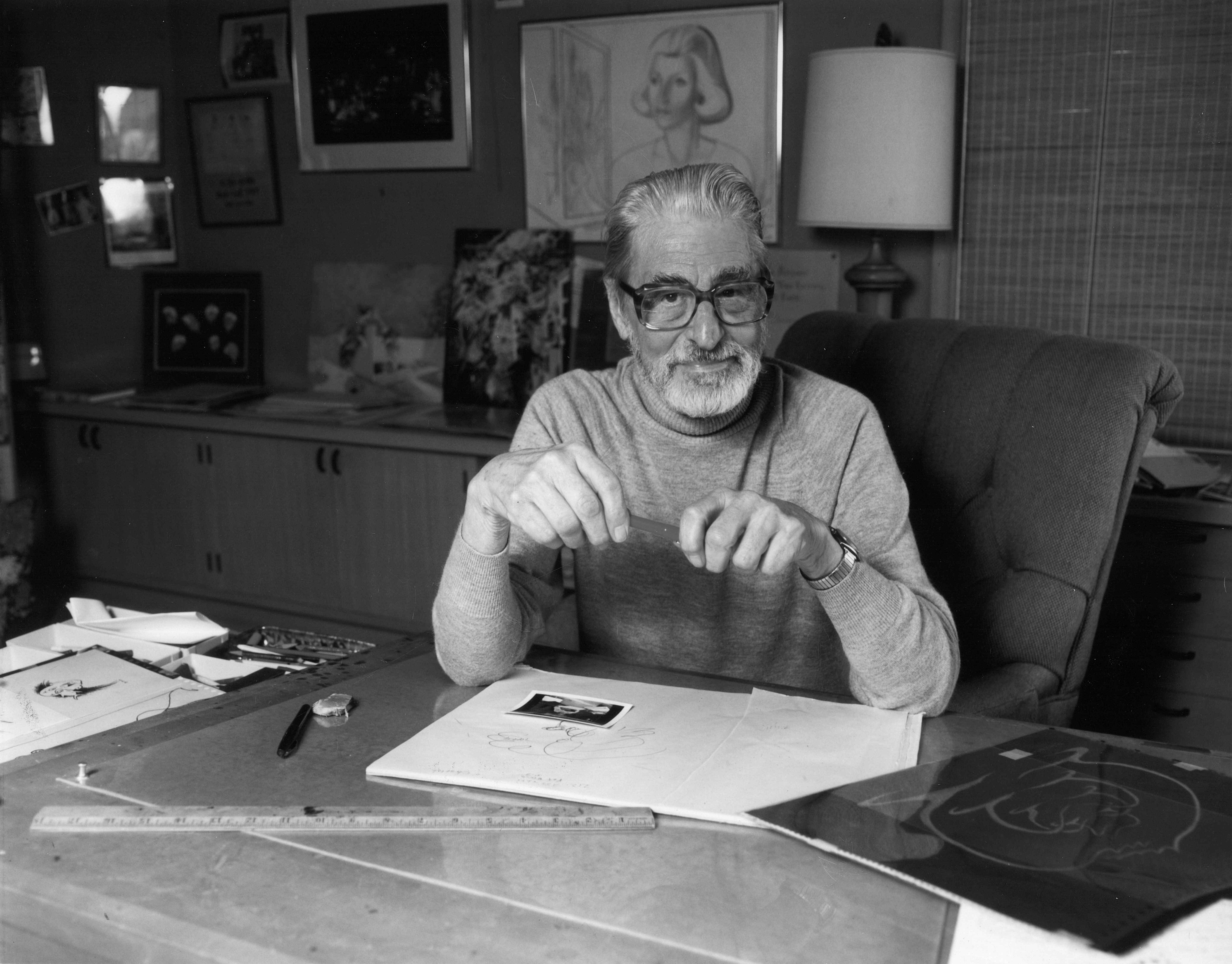 Theodor "Doctor Seuss" Geisel sits upright with a book on a desk in front of him. 